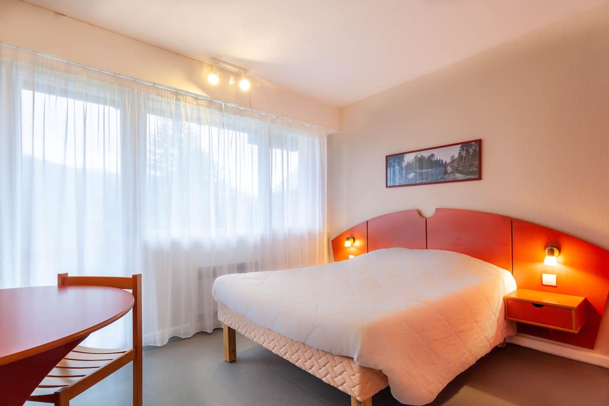 Una habitación de hotel con una cama, una mesita de noche con lámpara, una silla y una ventana con cortinas traslúcidas.