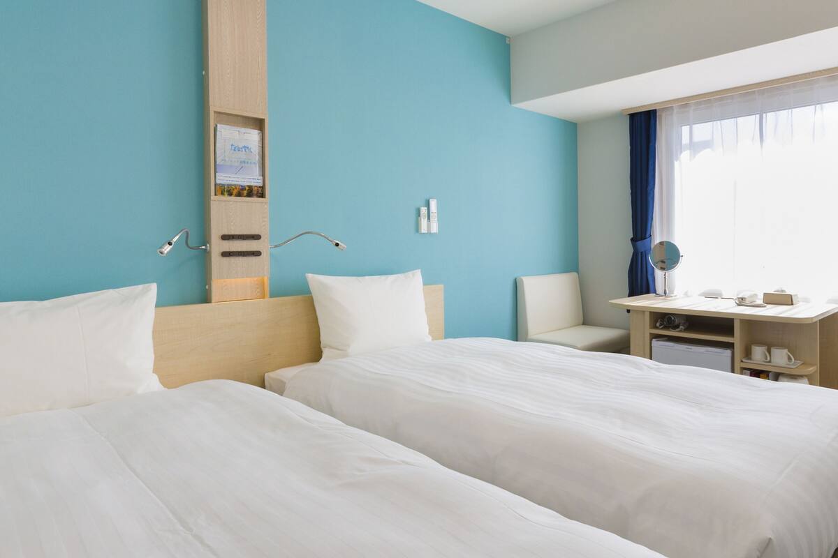Ein Hotelzimmer mit zwei Betten, einem Schreibtisch, einem Stuhl, einem Spiegel und einem Fenster mit Vorhängen.