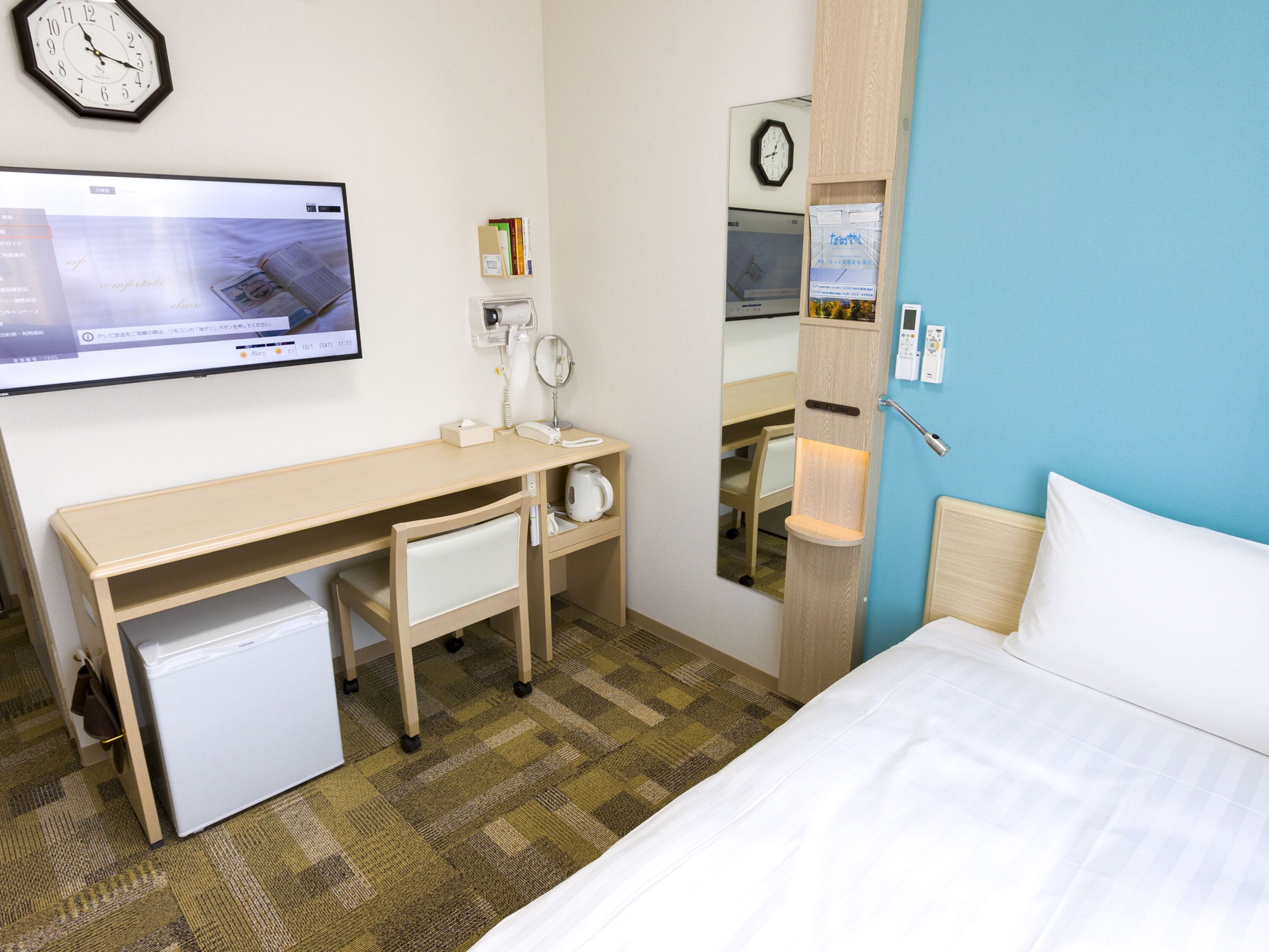 A hotel room with a bed, a desk with a computer, a small fridge, a mirror, and a clock on the wall.