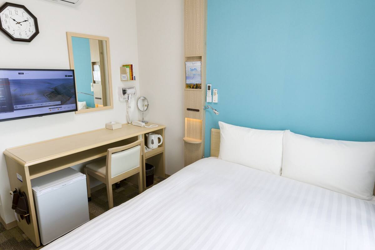 Ein Hotelzimmer mit Bett, Schreibtisch mit Computer, einer kleinen Kühlbox, einer Uhr und einem Spiegel.