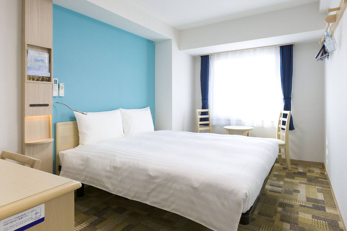 Ein Hotelzimmer mit Bett, Schreibtisch, Stuhl, Nachttisch und einem Fenster mit Vorhängen.
