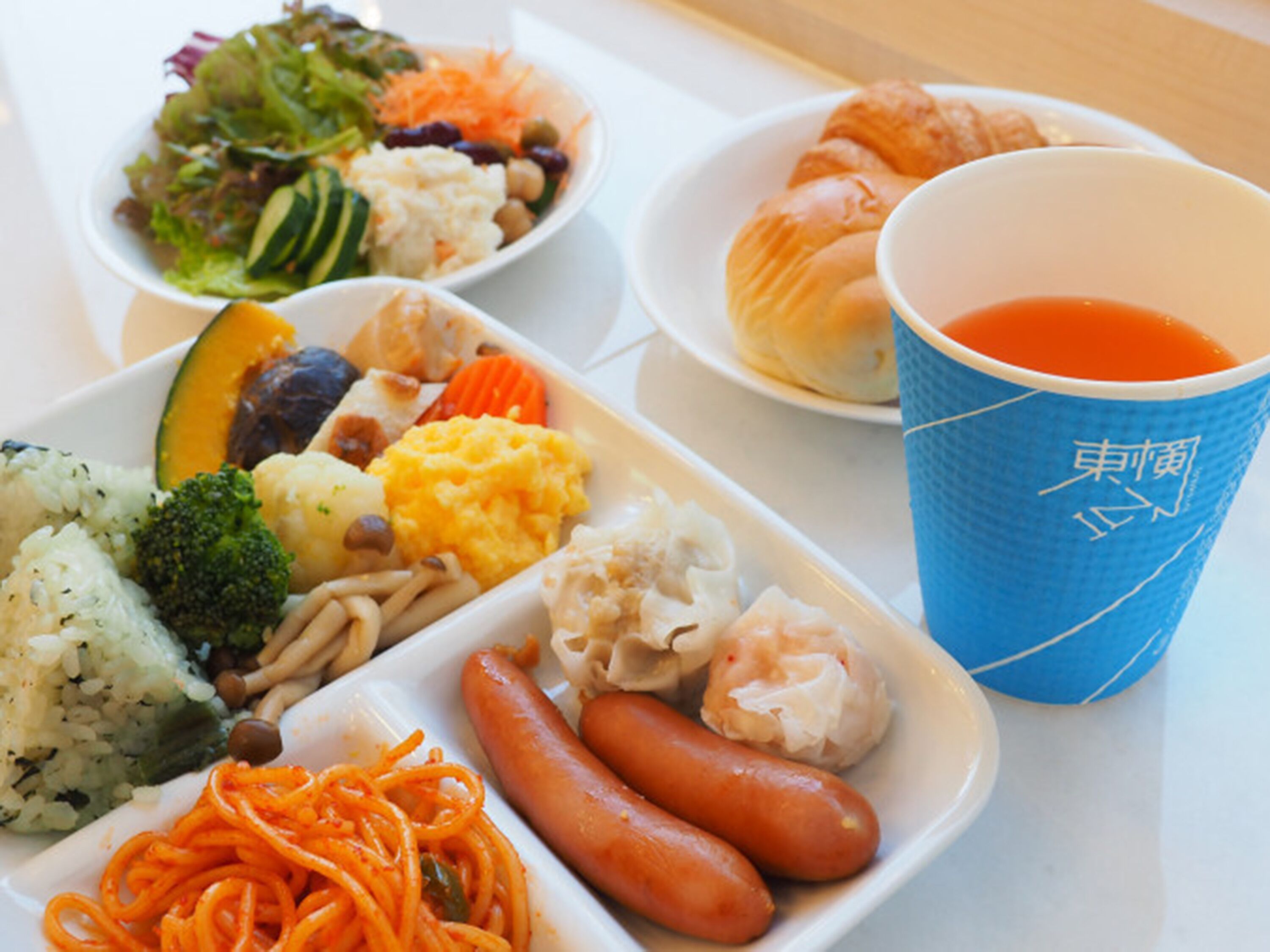 A meal with various dishes including pasta, vegetables, and sausages, along with a cup of tea.