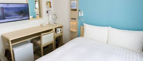 A hotel room with a bed, a desk with a computer, a small refrigerator, a mirror, and a clock on the wall.