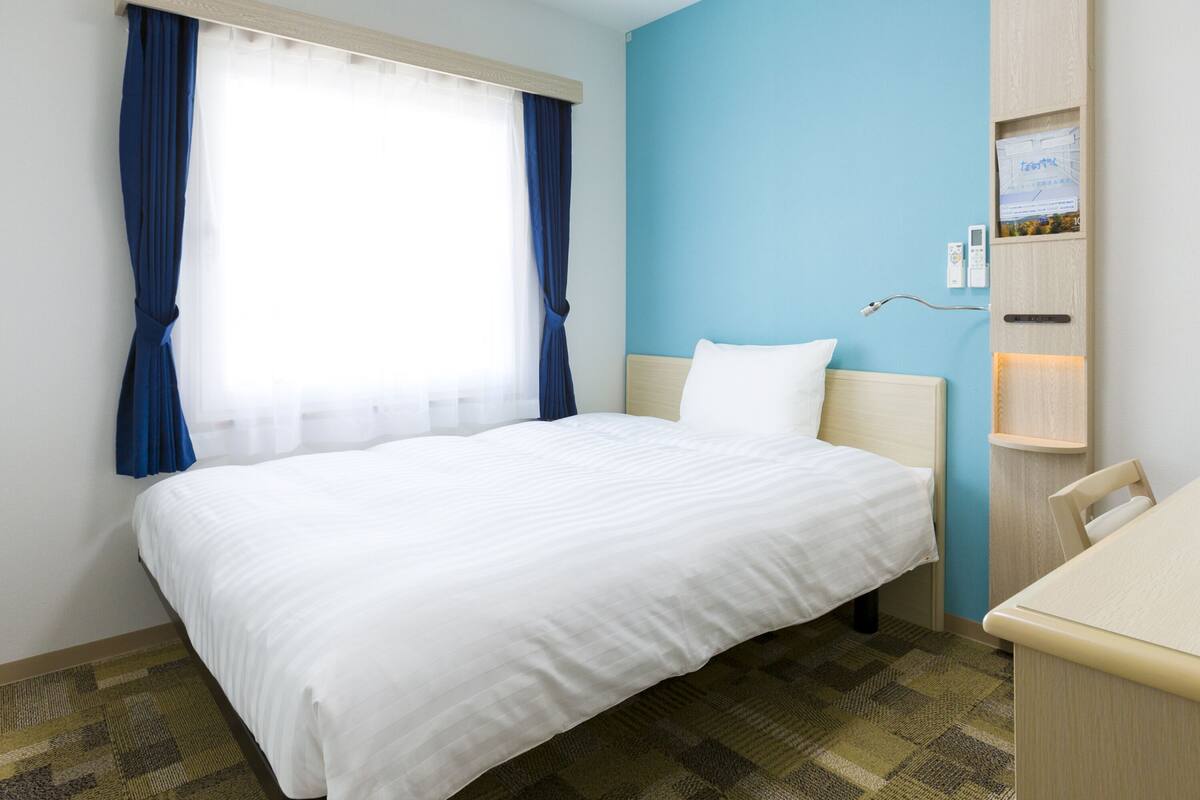 Ein Hotelzimmer mit Bett, Schreibtisch, einem Fenster mit Vorhängen, einem an der Wand montierten Fernseher und einer kleinen Ablage.