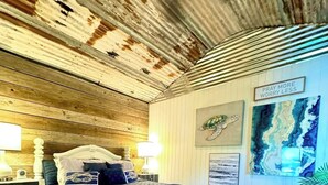 A bedroom with a wooden ceiling, a bed with a blue bedspread, two bedside lamps, and artwork on the walls.