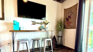 A cozy room with a wooden floor, a flat-screen TV mounted on the wall, a small bar area with stools, and a large window with curtains.