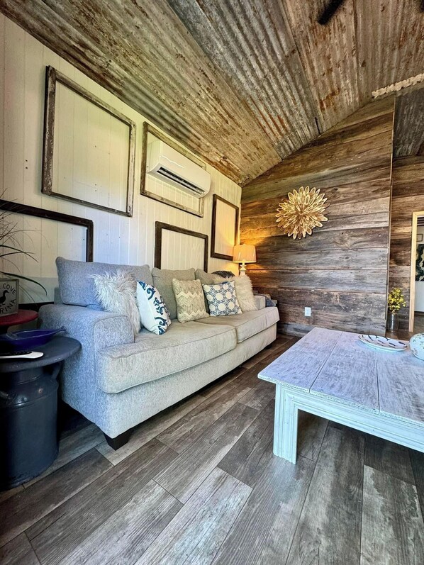 Un salón acogedor con techo de madera, un sofá gris con cojines estampados, una mesa de centro blanca y un aire acondicionado instalado en la pared.