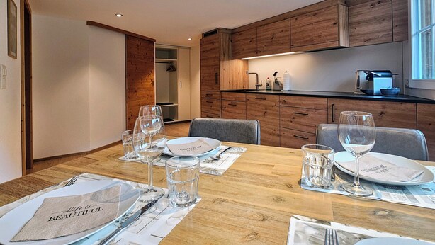 Une cuisine moderne avec des meubles en bois, une table à manger dressée pour quatre personnes, avec des verres et des assiettes, et une vue sur la pièce adjacente.