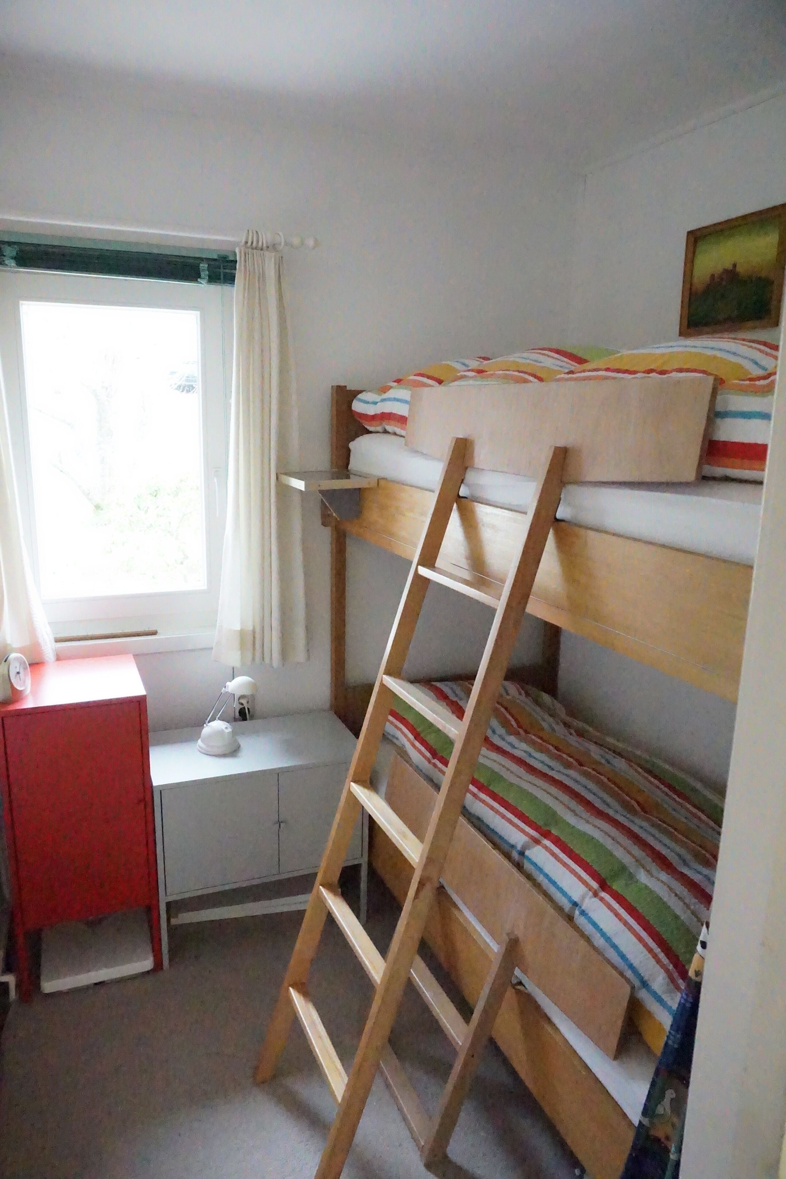 A bunk bed room with a ladder, a desk with a lamp, and a window with curtains.