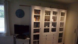 Una habitación con un mueble blanco de puertas de cristal que contiene libros y objetos decorativos, una pequeña mesa blanca con un televisor y un radiador.