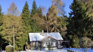 Ein Haus mit schneebedecktem Dach, umgeben von hohen immergrünen Bäumen und einem Holzzaun.