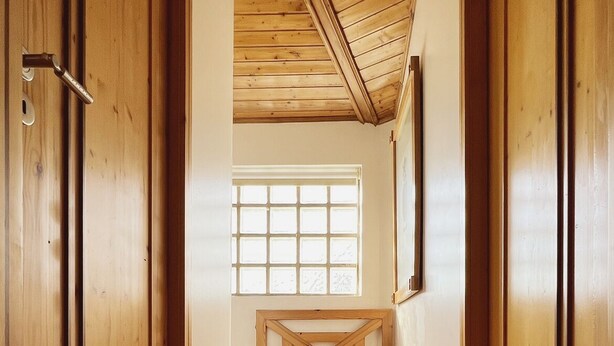Een houten gang met een hoge plafond, een raam aan het einde en een houten bankje.