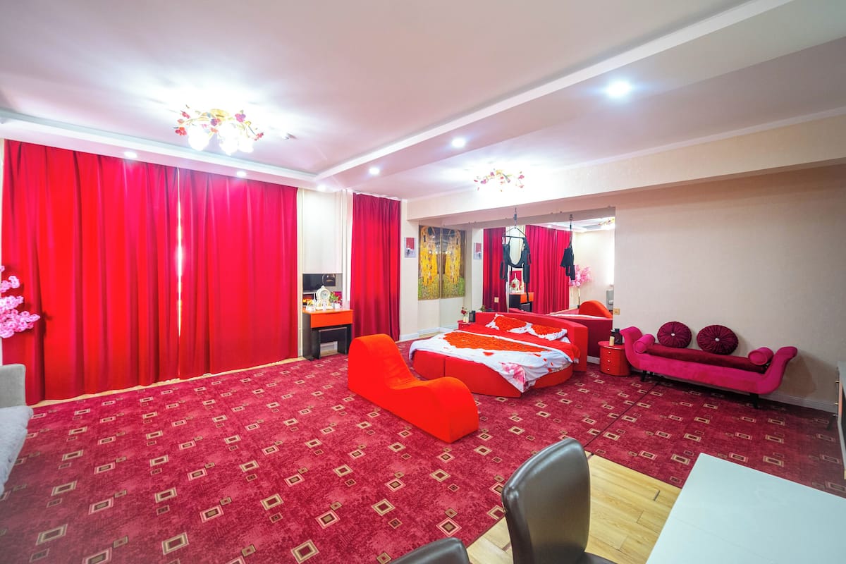 A hotel room with a red carpet, a bed with a floral bedspread, a red sofa, a desk with a chair, and a mirror.