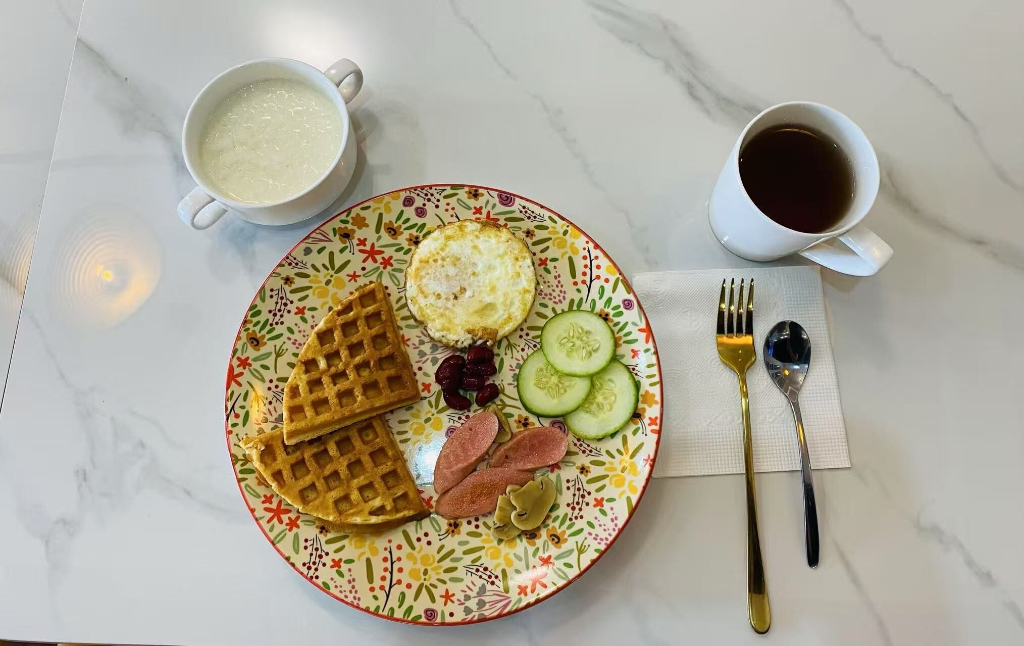 Une assiette contenant des gaufres, un œuf au plat, des rondelles de concombre et des saucisses, accompagnée d’une tasse de café et d’une tasse de thé, le tout posé sur une table en marbre.