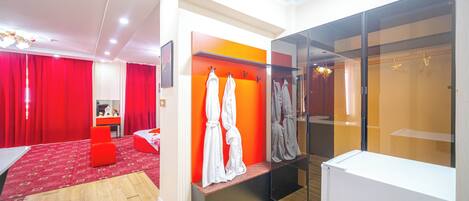 Une chambre d’hôtel moderne avec une moquette rouge, un plancher en bois, un lit, un coin salon, une armoire avec des vêtements suspendus et une cabine de douche en verre.