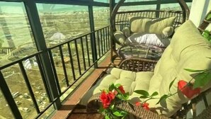 Ein Balkon mit Korbstuhl und Kissen, ein kleiner Tisch mit Blumen und Blick auf eine Landschaft durch große Fenster.