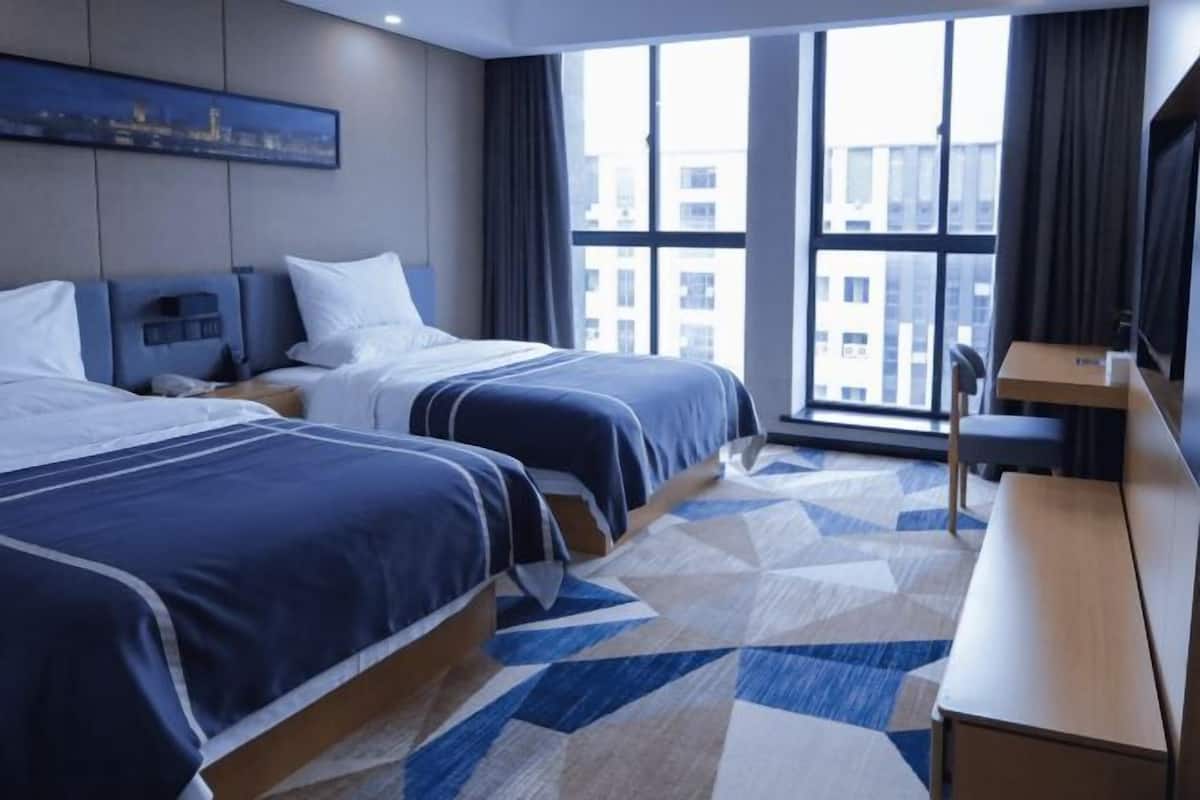 A hotel room with two beds, a desk, and a large window offering a city view.