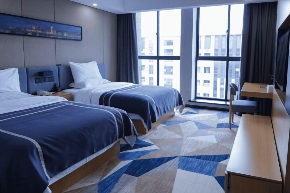 A hotel room with two beds, a desk, a large window with a city view, and a geometric patterned carpet.