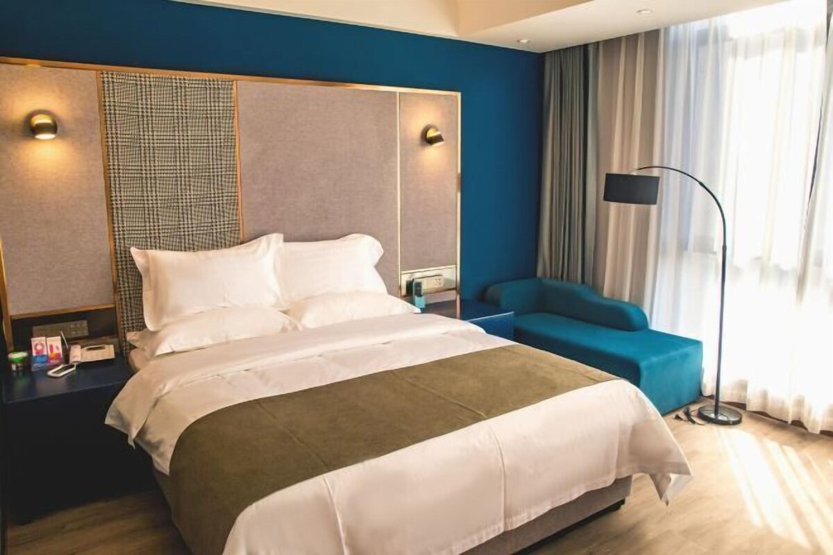 A hotel room with a large bed, a blue armchair, a bedside table with a lamp, and a view of the outdoors through sheer curtains.