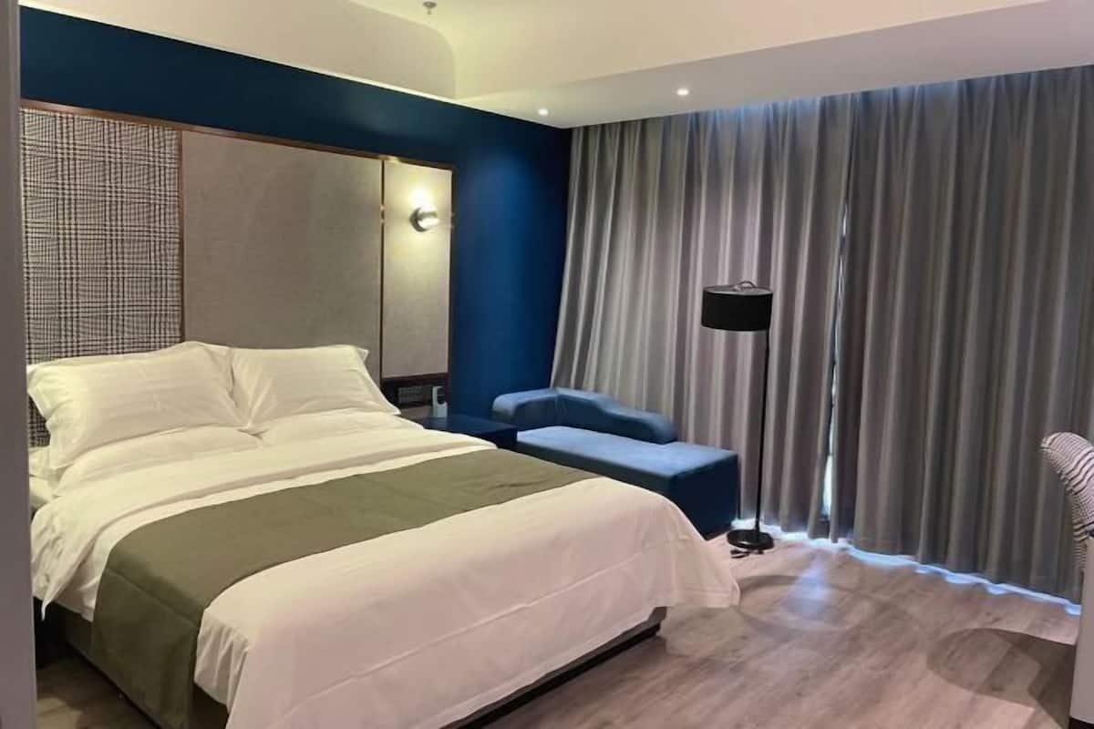 A modern hotel room with a large bed, a seating area, a desk, and a window with curtains.