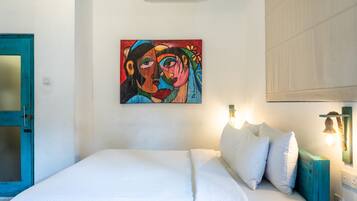 Un dormitorio con una cama, ropa de cama blanca, un cuadro de colores en la pared y una puerta azul.