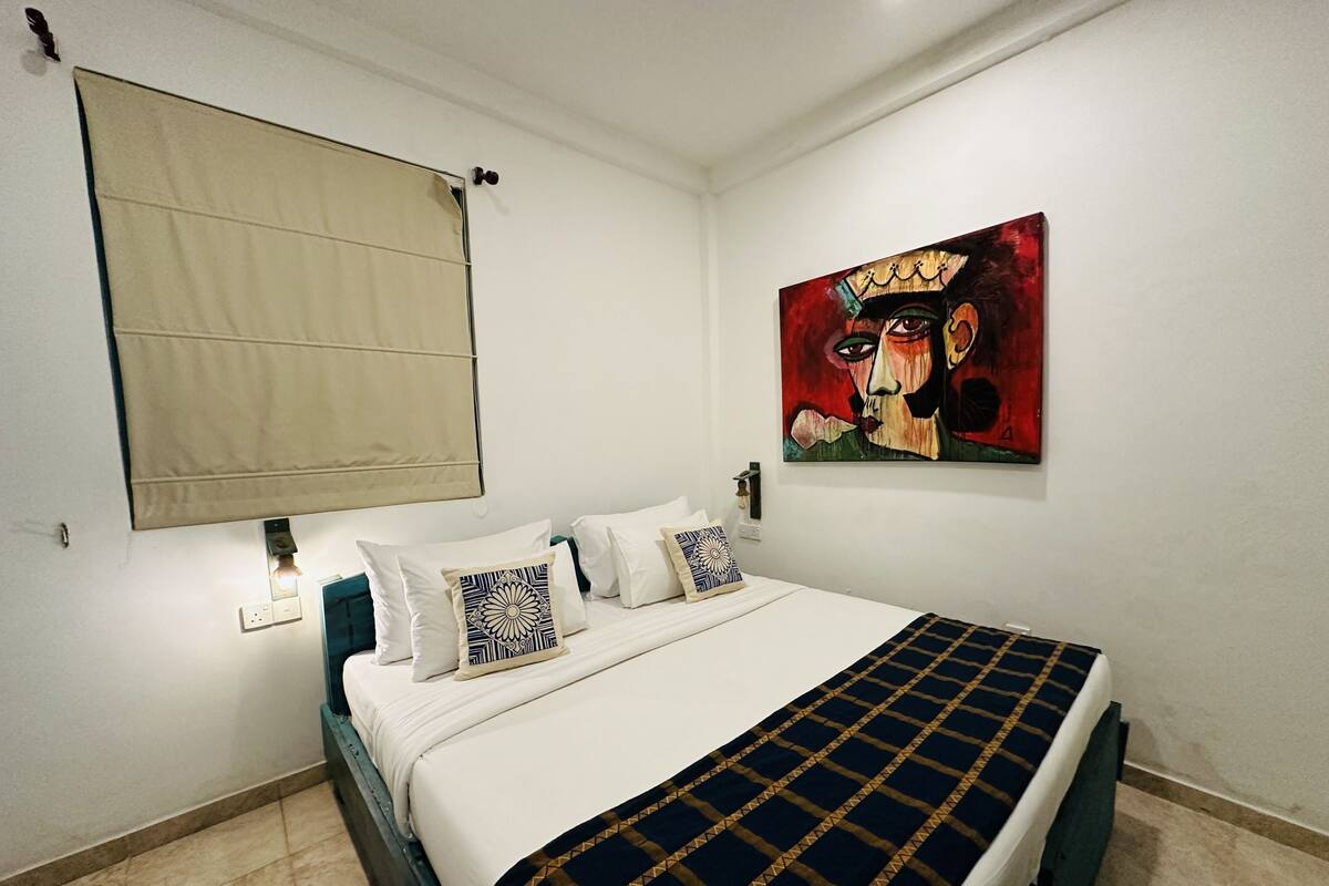 Een slaapkamer met een bed, kussens, een schilderij en een raam met een rolluik.