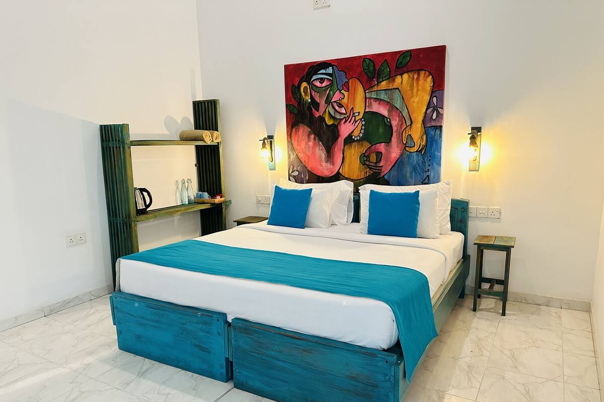 Een moderne slaapkamer met een groot bed, blauwgroene beddengoed, een kleurrijk abstract schilderij, nachtlampjes en een klein houten nachtkastje.