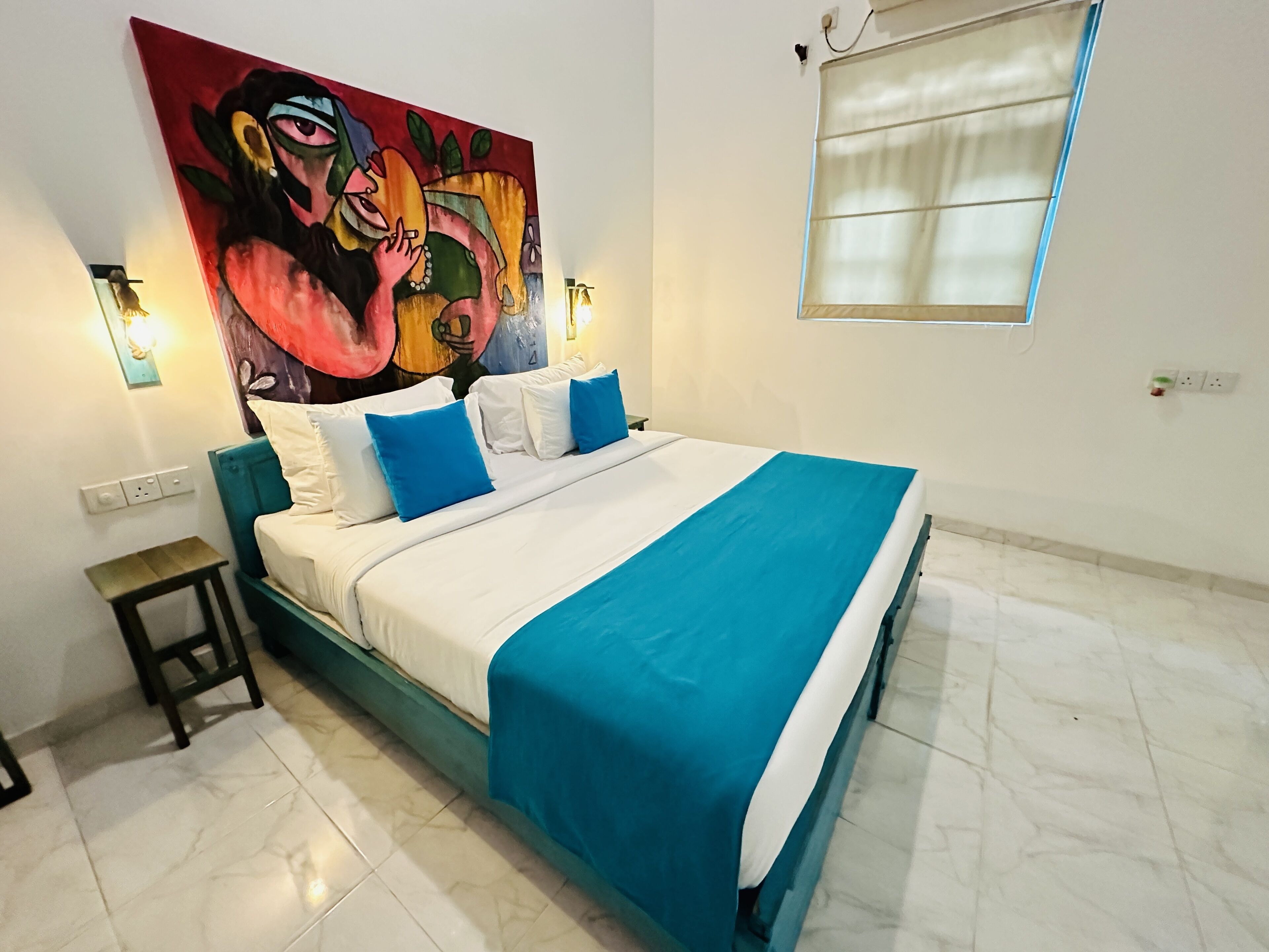 A hotel room with a large bed, white and blue bedding, a teal headboard, a small bedside table, and a colorful abstract painting on the wall.