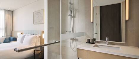 A modern hotel room with a glass-enclosed shower, a large bed with white linens, a wooden headboard, and a large mirror above the sink.