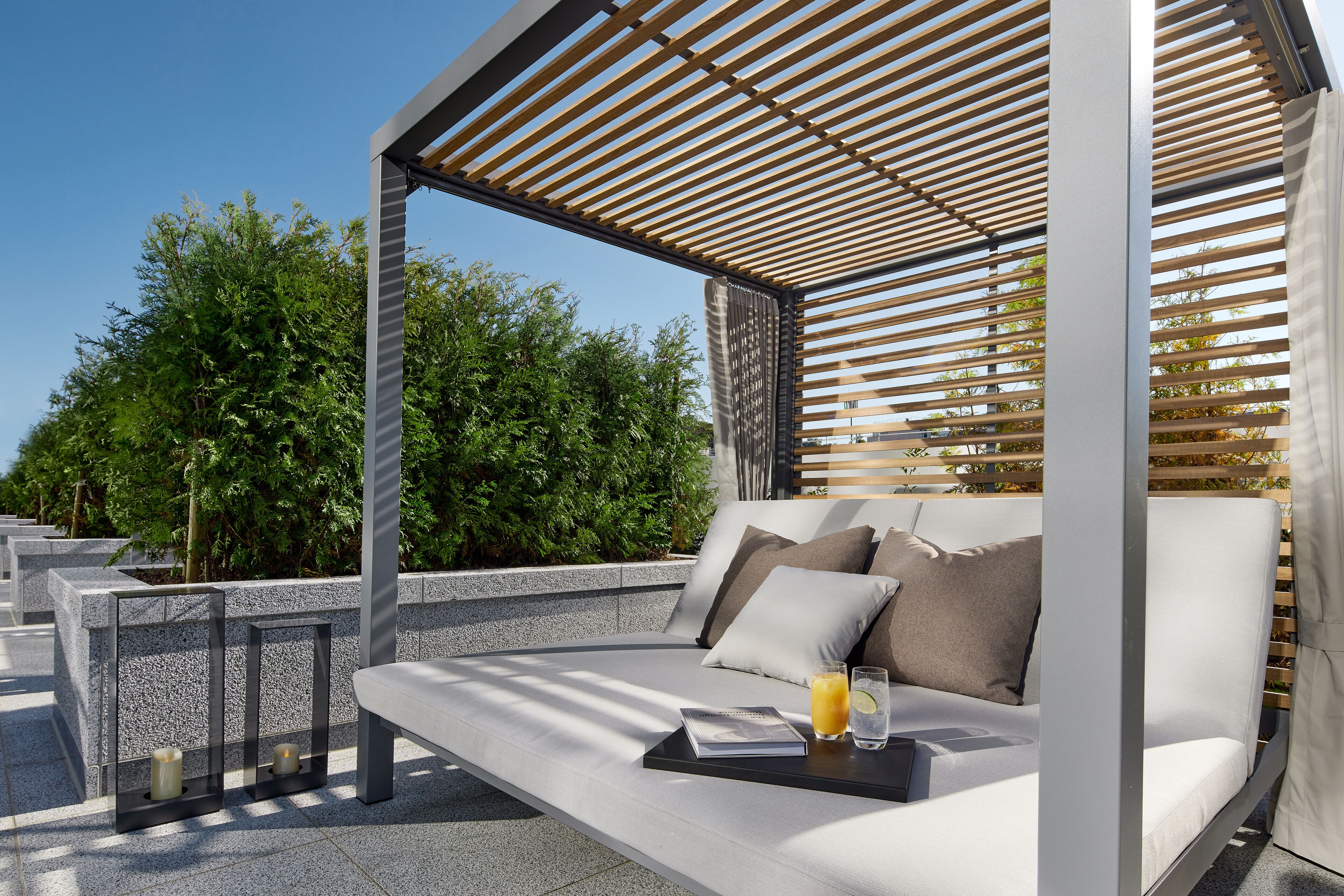 A modern outdoor seating area with a cushioned bench, a glass table with a drink, and a wooden pergola structure.
