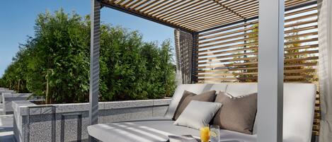 Un espacio exterior moderno con un banco acolchado, una mesa de vidrio con una bebida y una estructura de pérgola de madera.