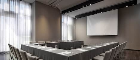 Un salón de conferencias con una larga mesa, sillas, una pantalla de proyector y un amplio ventanal con cortinas.
