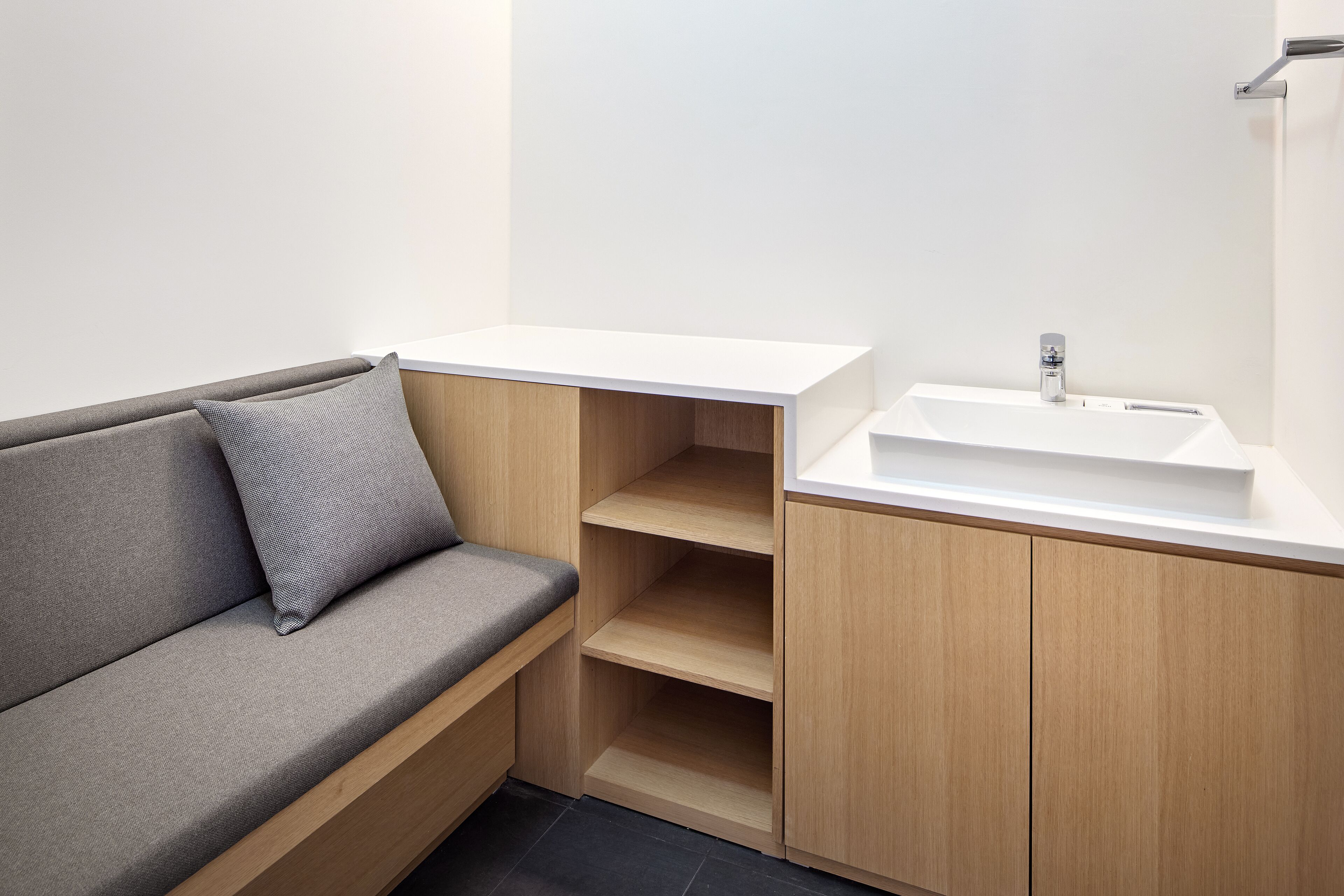 Een moderne badkamer met een houten wastafelmeubel, een witte wasbak en een grijze bank met een kussen.
