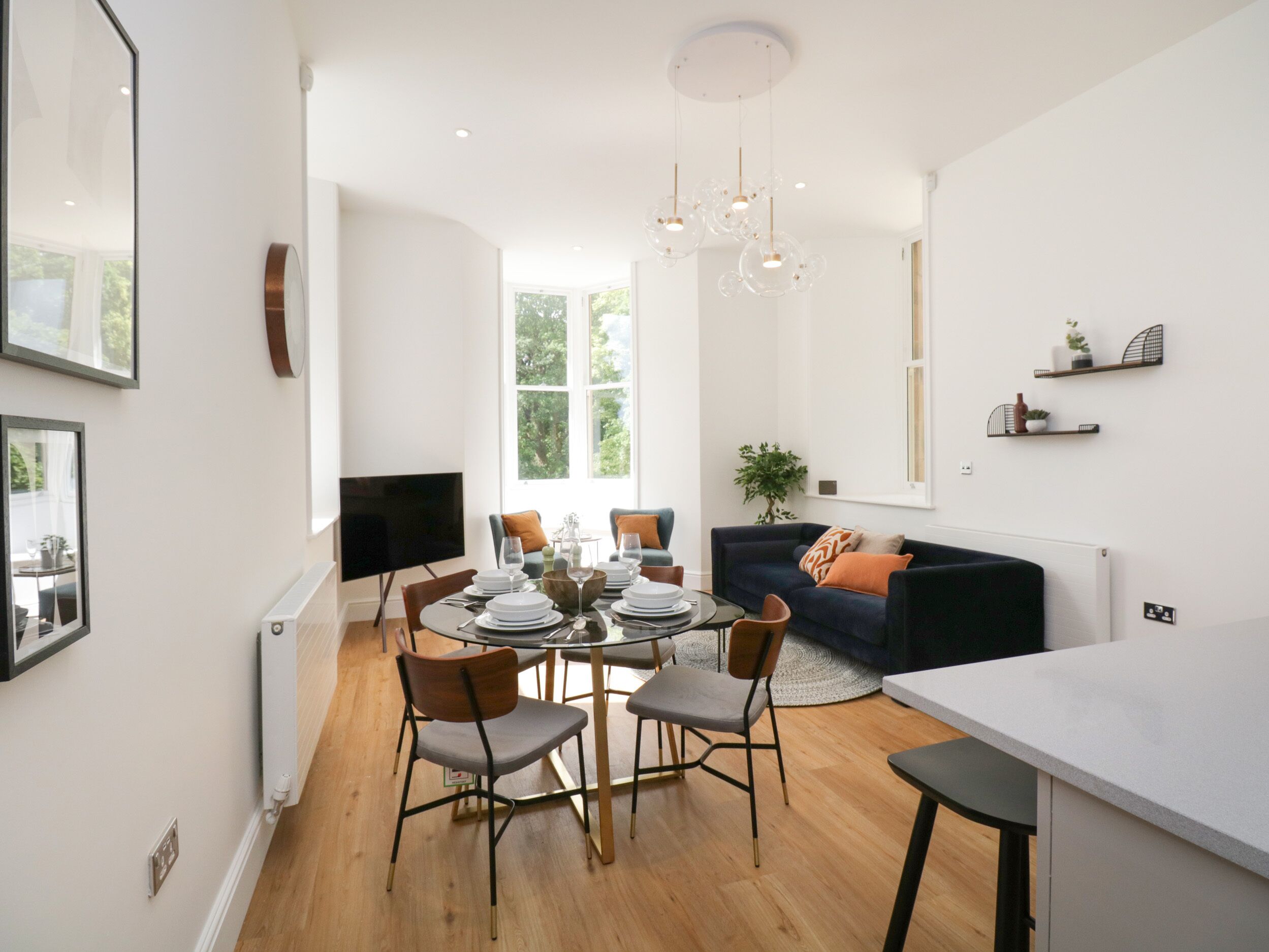 A modern living room with a dining area, featuring a round table set for four, a dark blue sofa, and a large window providing natural light.