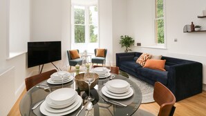 A modern living room with a round glass table set for dining, a dark blue sofa with orange cushions, and a large window providing natural light. - The Florence Suite Stone Cross Mansion (Ulverston)