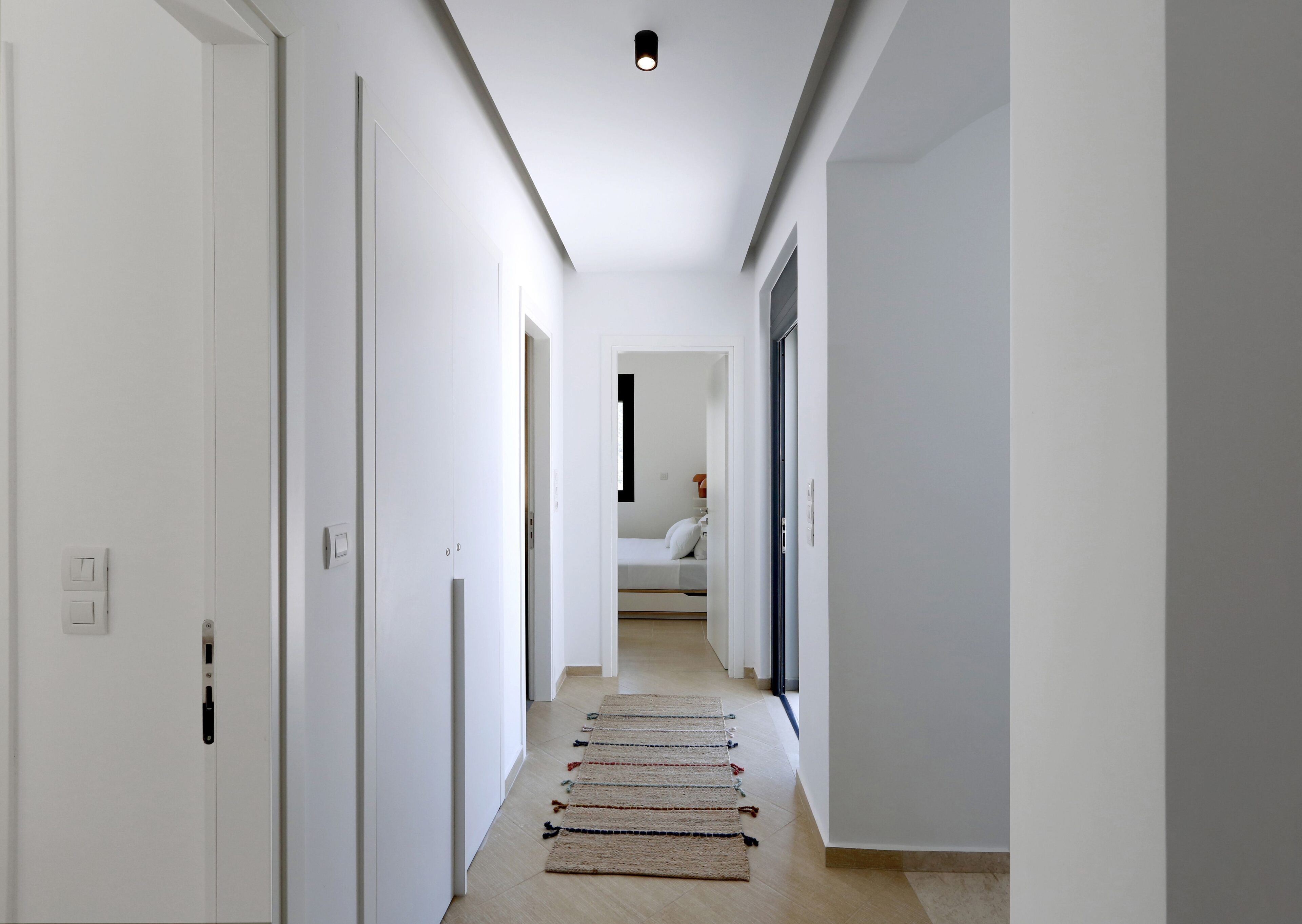 A hallway with white walls, a wooden floor, and a bed in the distance.