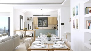 A modern dining area with a wooden table set for six, white chairs, and a view of the kitchen in the background. - Aires Villa (Agios Nikolaos)