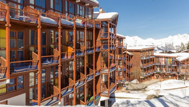 Un hôtel à plusieurs étages doté de balcons, de grandes fenêtres et surplombant un paysage montagneux enneigé.