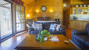 A cozy living room with a wooden ceiling, a sofa set, a coffee table with a plant, and a kitchen area with cabinets and a microwave. - Silky Oak Cottage & Studio - Delightful country cottage along popular Hermitage Rd (Pokolbin)