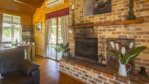 A cozy living room with a brick fireplace, wooden ceiling, and a dining area with a table and chairs. - Silky Oak Cottage & Studio - Delightful country cottage along popular Hermitage Rd (Pokolbin)