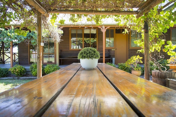 Une table de pique-nique en bois, ornée d’une plante en pot au centre, entourée d’une tonnelle en bois et d’un petit jardin planté de diverses espèces, le tout délimité par une clôture en bois.