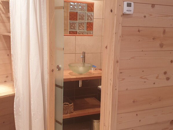 A wooden cabin bathroom with a glass bowl sink, a wooden shelf, and a tiled backsplash.