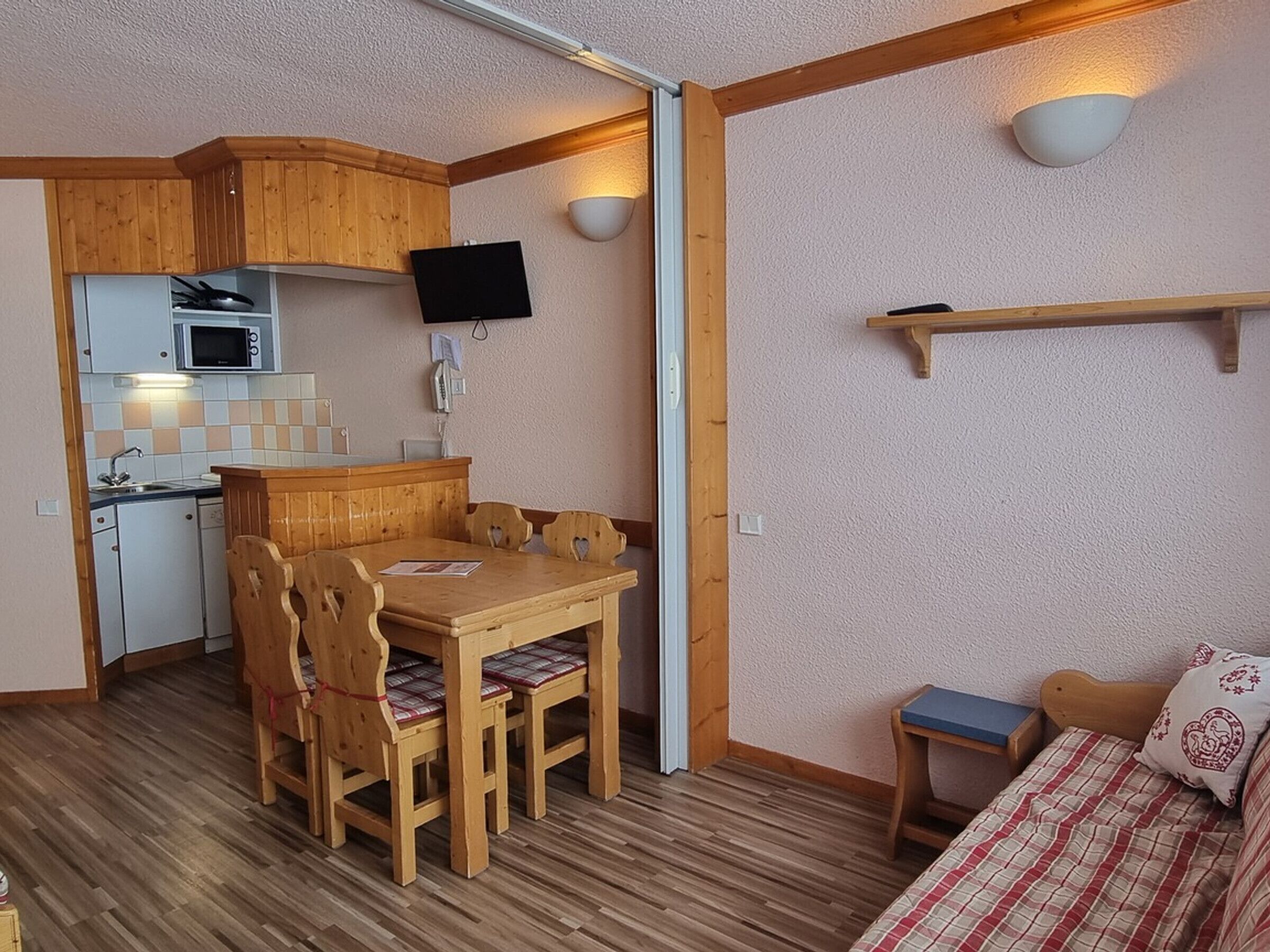 Habitación pequeña con una mesa y sillas de madera, una kitchenette compacta, una cama con cubrecama a cuadros y un televisor montado.