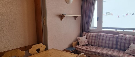 Une pièce chaleureuse avec une table en bois, des chaises, un canapé et une fenêtre donnant sur une plage où l’on peut voir des gens.