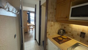 A compact kitchen with a microwave, sink, and a small stove. There is a wooden cabinet above the stove and a tiled backsplash. A chair and table are visible in the adjacent room. - Charming Ski-In Studio · Pets Welcome (Plagne Aime 2000)