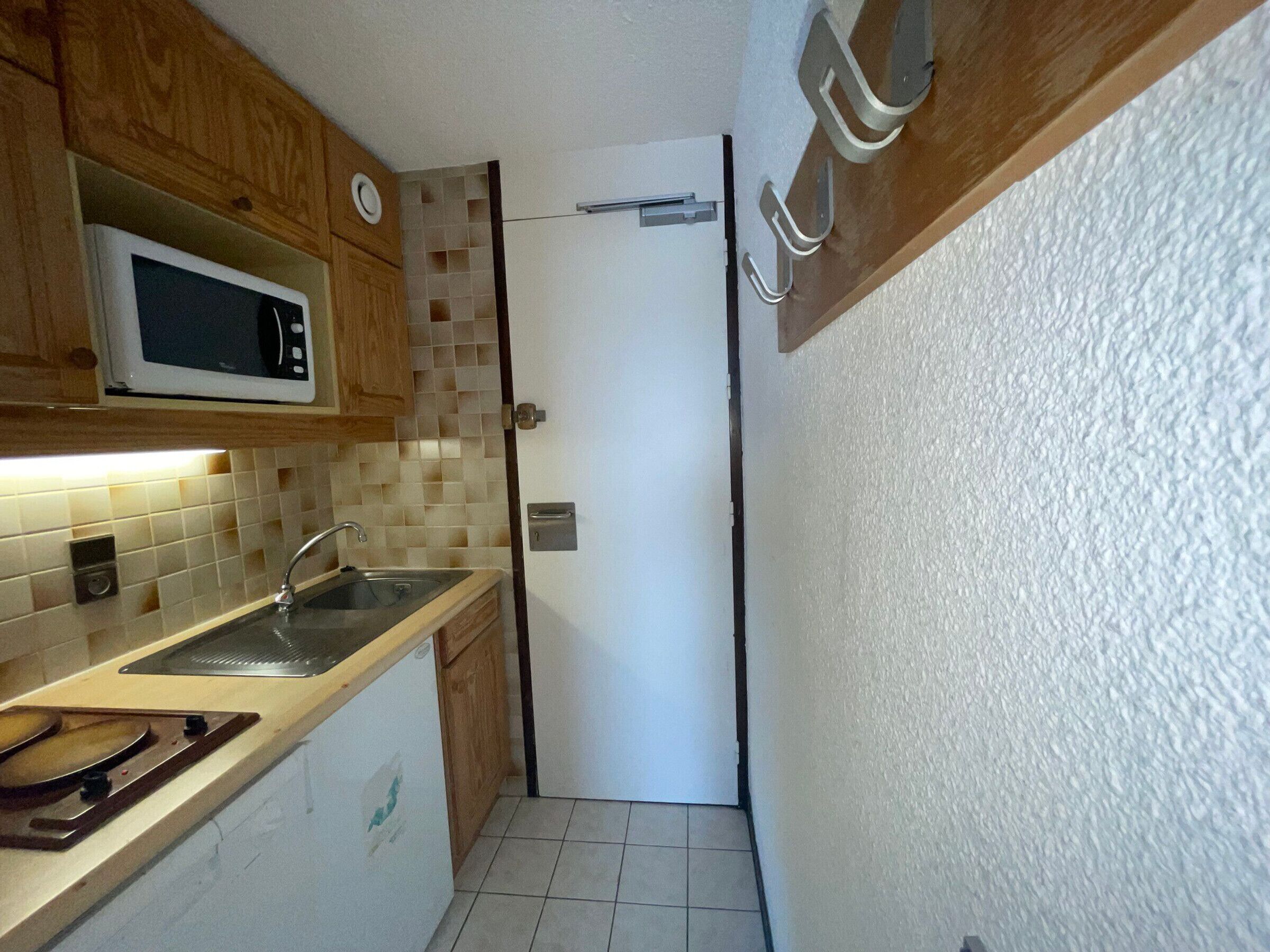 A compact kitchen with wooden cabinets, a microwave, a sink, and a door leading to another room.