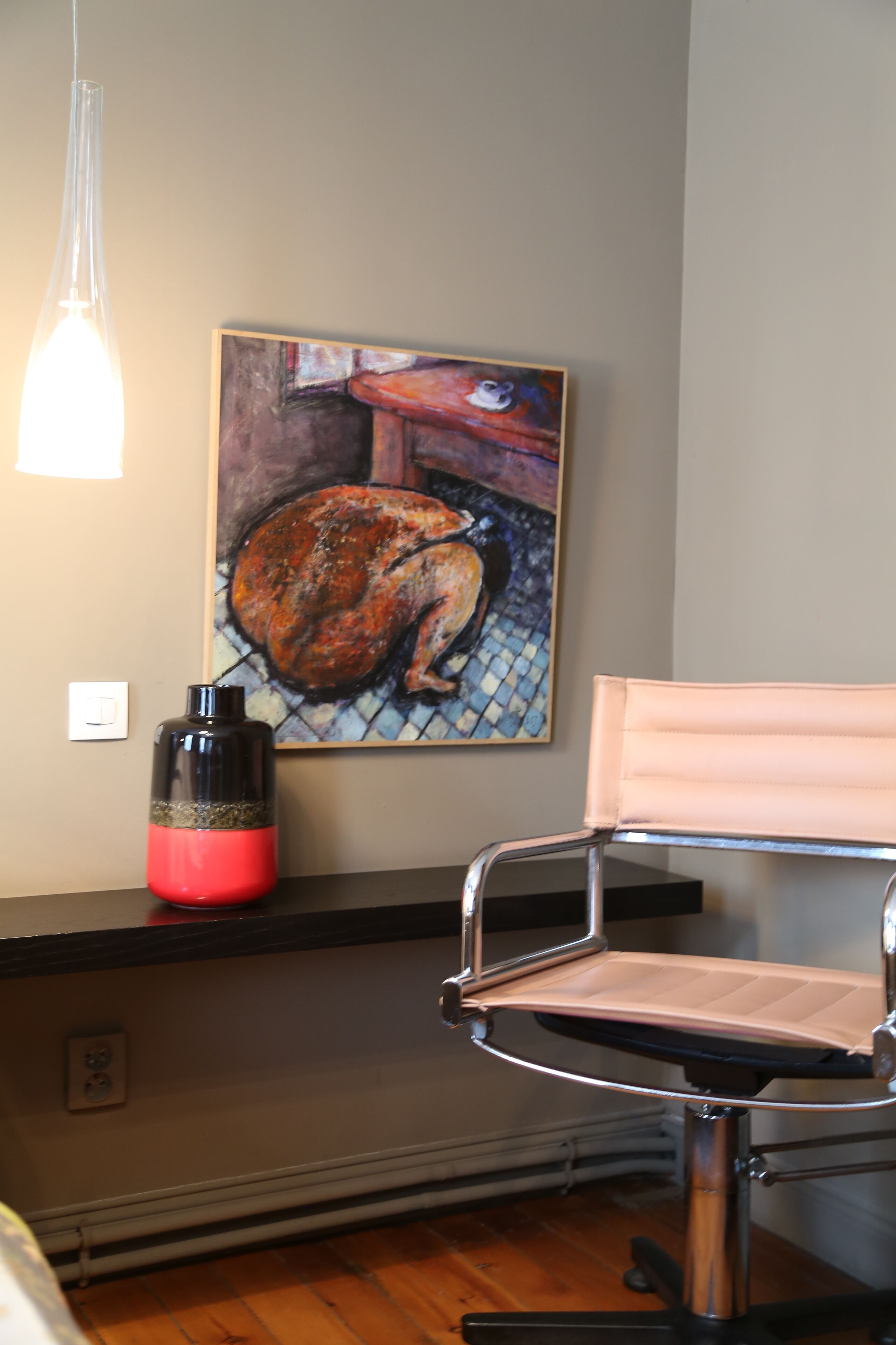 Una habitación con una silla, un cuadro, un objeto rojo y negro en una estantería, y una lámpara.