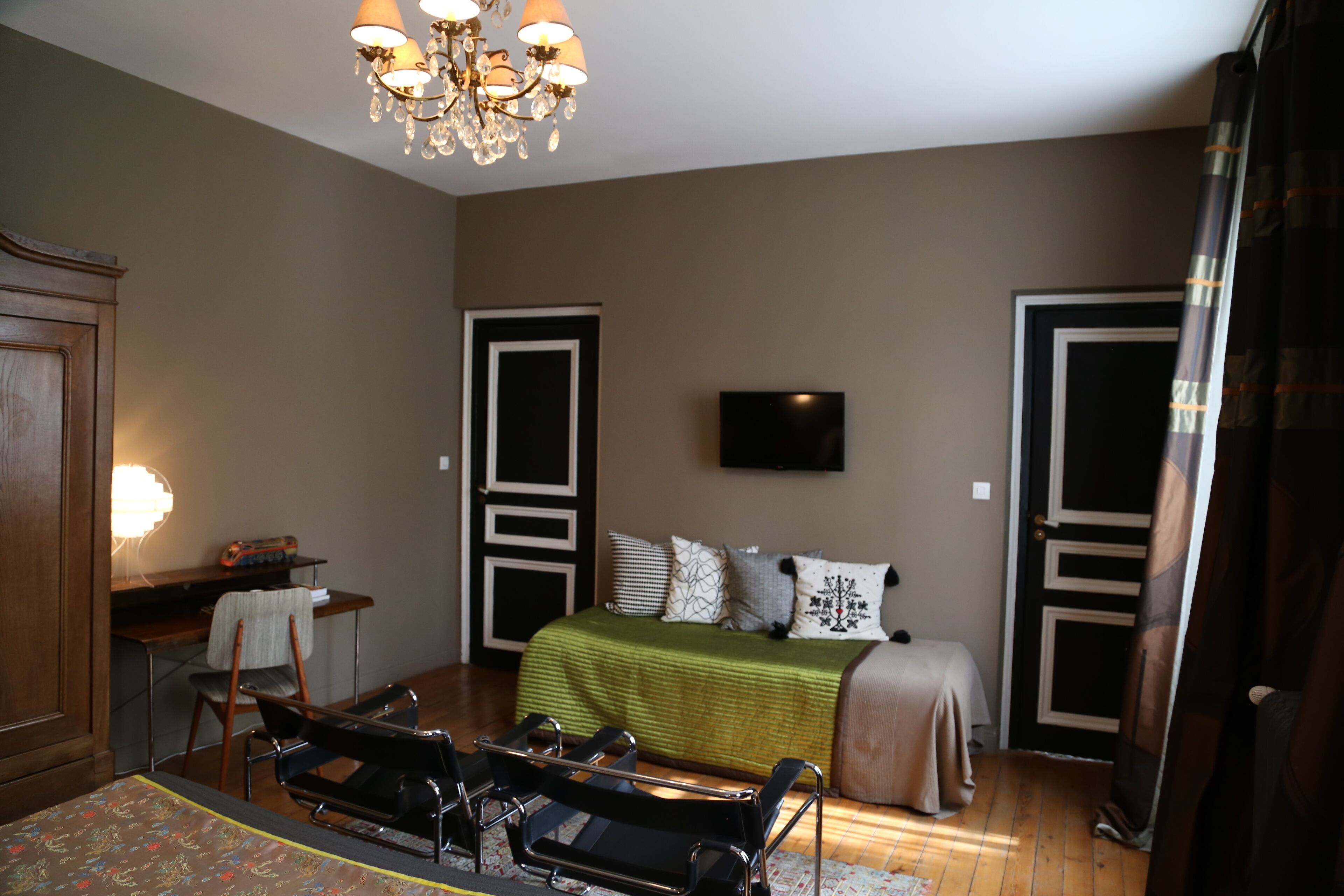 A bedroom with a bed, a desk, a chair, a television, and a chandelier.