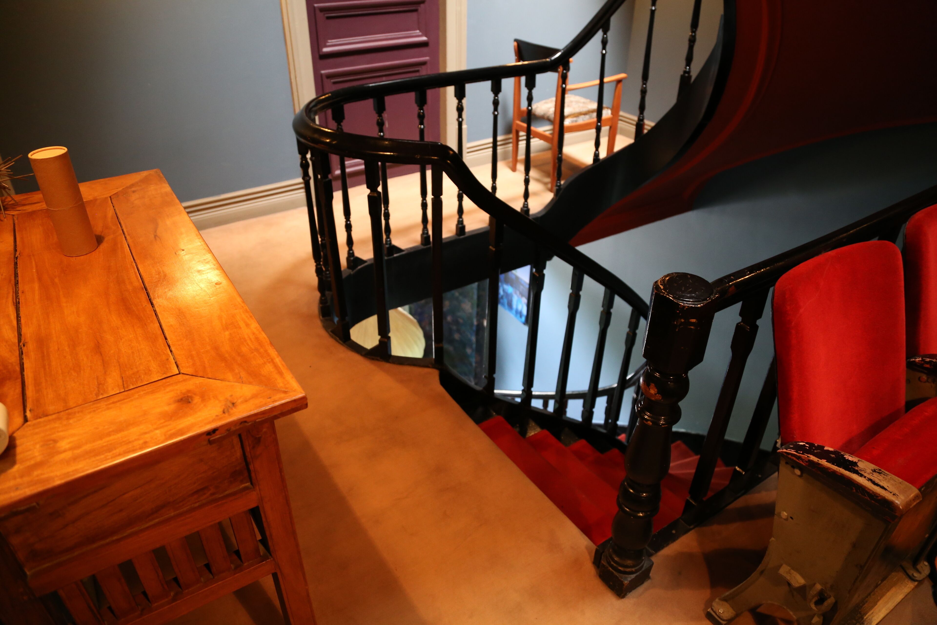 A wooden table with a cylindrical object on it, a black spiral staircase with red steps, and a red upholstered chair.