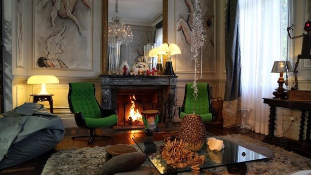 A cozy living room with a fireplace, green armchairs, a glass coffee table, and a fireplace mantle with decorative items.